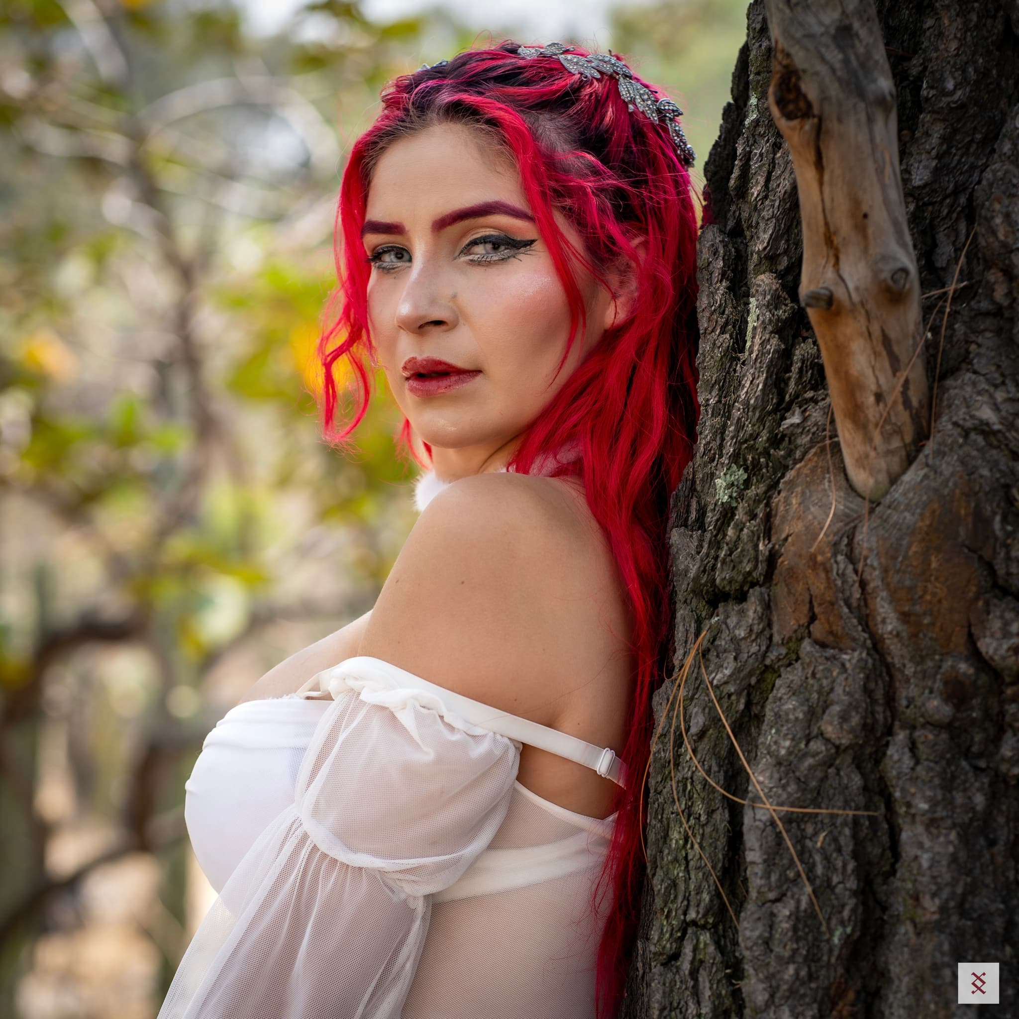 A person with vibrant red hair and dramatic makeup stands against a tree, wearing an off-the-shoulder white top, in a natural outdoor setting