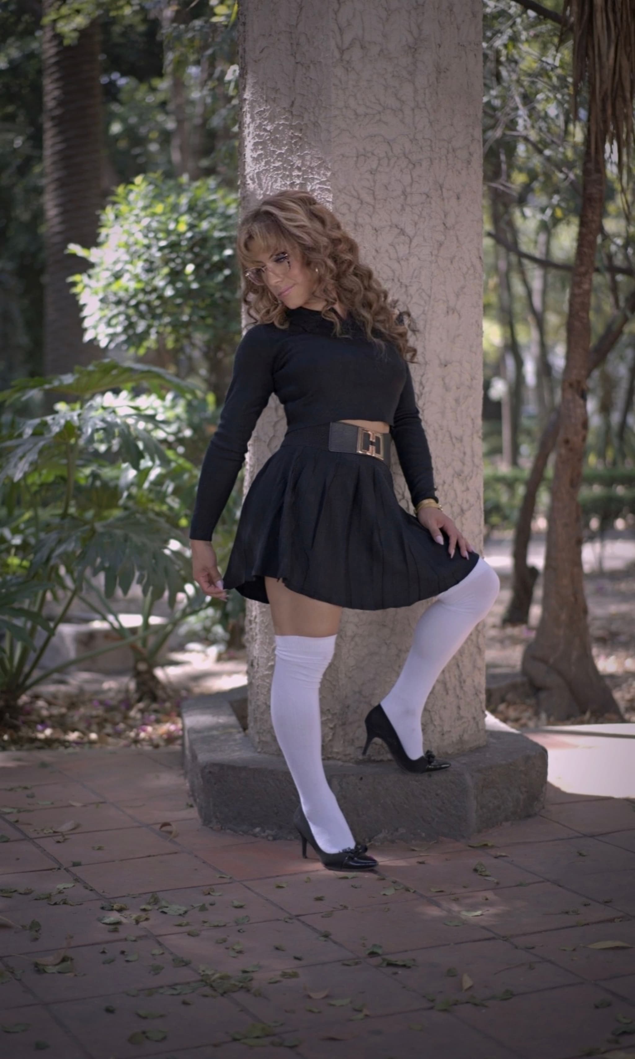 A person in a black dress and white knee-high socks poses outdoors against a tree trunk, surrounded by greenery