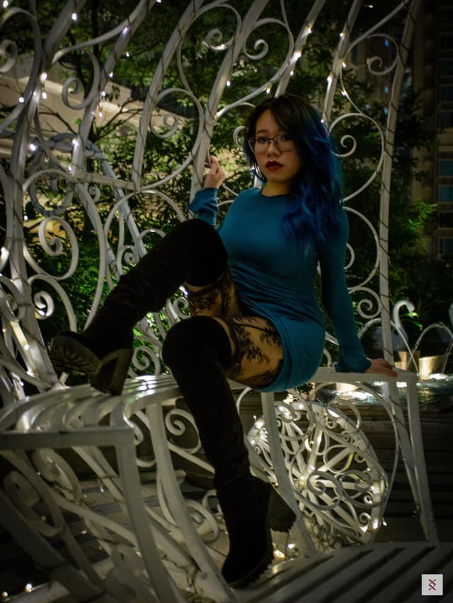 A person with blue hair and glasses poses on an ornate, white metal structure adorned with lights, wearing a blue dress and black thigh-high boots