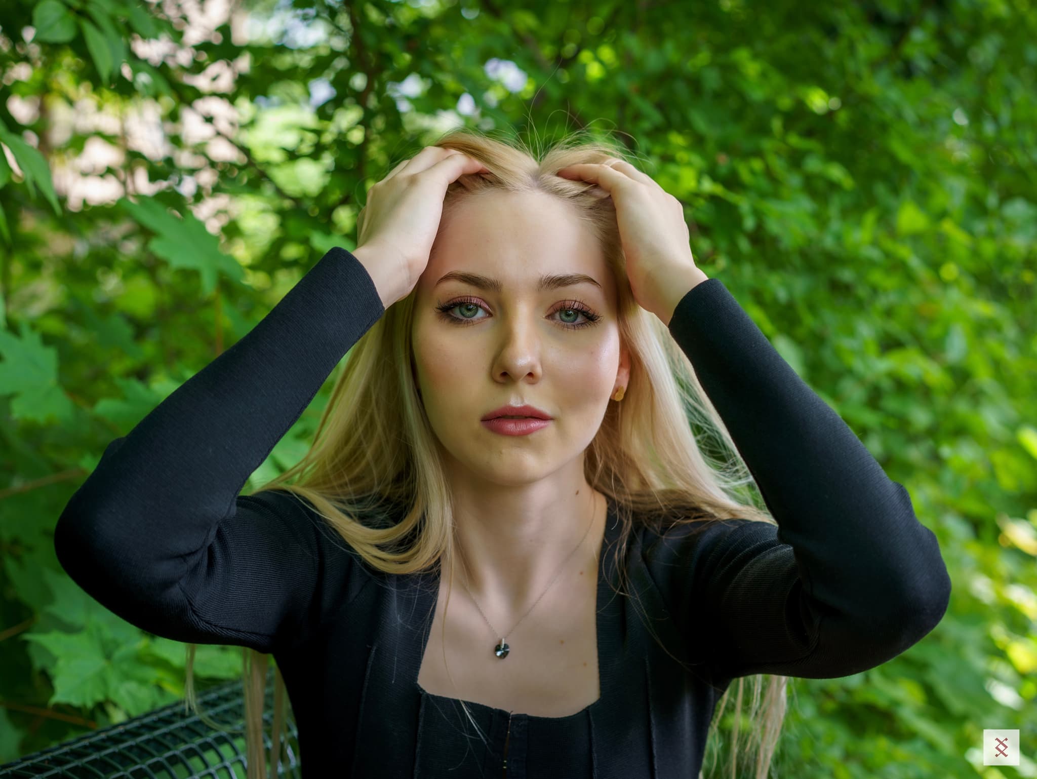 A person with long blonde hair, wearing a black top, is holding their head with both hands while standing in a lush green outdoor setting