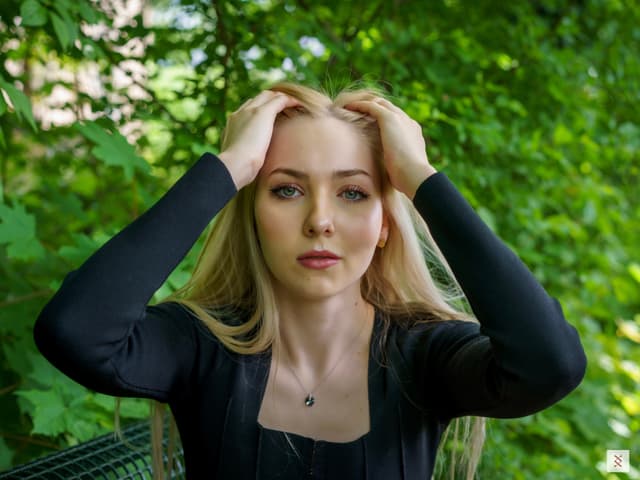 A person with long blonde hair, wearing a black top, is holding their head with both hands while standing in a lush green outdoor setting